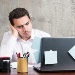 Homme assis a un bureau, l air agace devant l ecran de son ordinateur portable